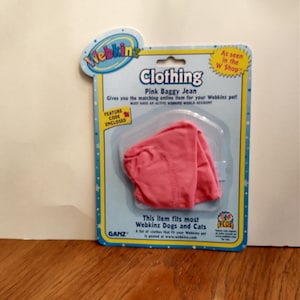 May include: A packet of pink baggy jeans for Webkinz pets. The packaging is blue with white stars and the Webkinz logo. The text "Clothing" and "Pink Baggy Jean" are visible on the front.