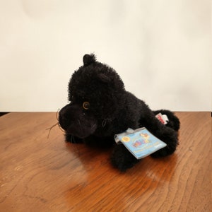 Peut inclure: Jouet en peluche de chat noir avec une étiquette Webkinz. Le chat est couché sur le côté, la tête tournée vers la droite. L'étiquette est bleue avec du texte blanc et une image d'un chat.