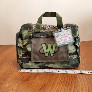 May include: A camouflage green plush pet carrier with a mesh top and a zipped closure. The carrier has a green W logo and a tag that reads "Plush Pet Carrier".