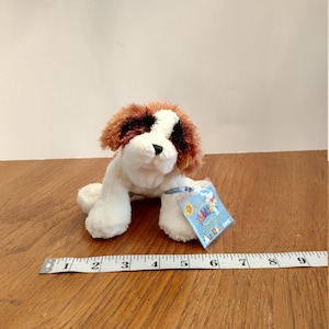 May include: White, brown and black plush dog toy with a blue tag that says "Webkinz Signature Ganz". The toy is approximately 5 inches long and is sitting on a wooden surface.