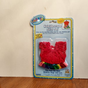 May include: Webkinz Kaleidescope Skirt Set for dogs and cats. The set includes a red top with a rainbow skirt. The packaging features the Webkinz logo and a plus sign.