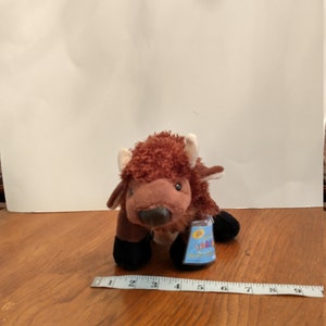 May include: A brown and white stuffed bison with black hooves and a Webkinz tag. The bison is sitting on a wooden surface.
