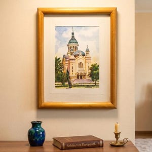 Puede incluir: Una pintura en acuarela enmarcada de una catedral con cúpula verde, en un marco de madera clara. Debajo del marco hay un jarrón de cerámica azul y verde, un libro marrón titulado Arhitectura Europeană y un pequeño candelabro de latón con vela.