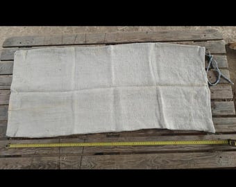 May include: A rectangular, off-white burlap sack with a blue rope attached to one end. A yellow measuring tape, marked in inches, lies along the bottom edge of the sack, indicating its length. The sack is placed on a wooden pallet.