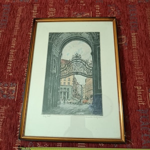 Puede incluir: Un grabado enmarcado que representa un arco de hierro forjado ornamentado que enmarca una escena de calle con edificios y vehículos. Una cinta métrica amarilla está colocada en la parte inferior, mostrando que el ancho es de 33 centímetros.