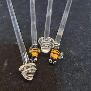 May include: Four clear glass stir sticks with decorative ends. Two sticks have a bee design with yellow and black stripes. The other two have a swirl and a round design. The sticks are on a dark surface.