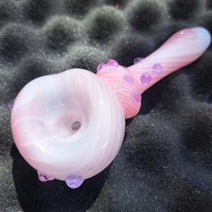 May include: A pink and white glass pipe with a swirl design. The pipe has a bowl, a stem, and a mouthpiece. The bowl has a large opening, and the stem is decorated with small, clear, pink glass beads. The mouthpiece is a light blue color.