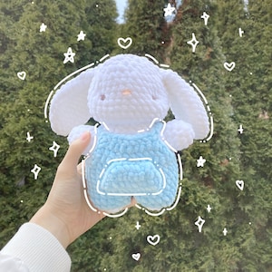 May include: A white crocheted bunny wearing blue overalls with a pocket. The bunny is being held in a hand against a green background.