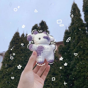 May include: A crocheted white and purple cow plush toy with black eyes and a pink nose. The toy is being held in a hand against a background of evergreen trees.