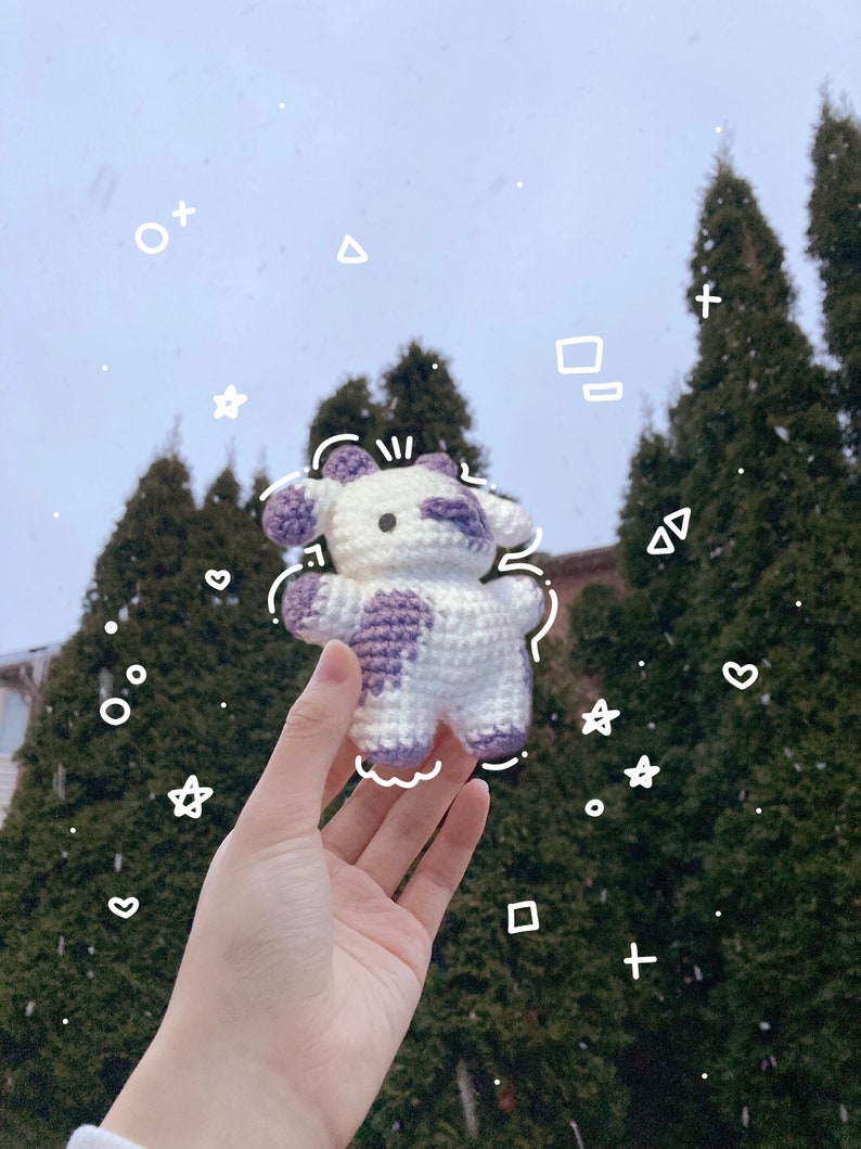 May include: A crocheted white and purple cow plush toy with black eyes and a pink nose. The toy is being held in a hand against a background of evergreen trees.