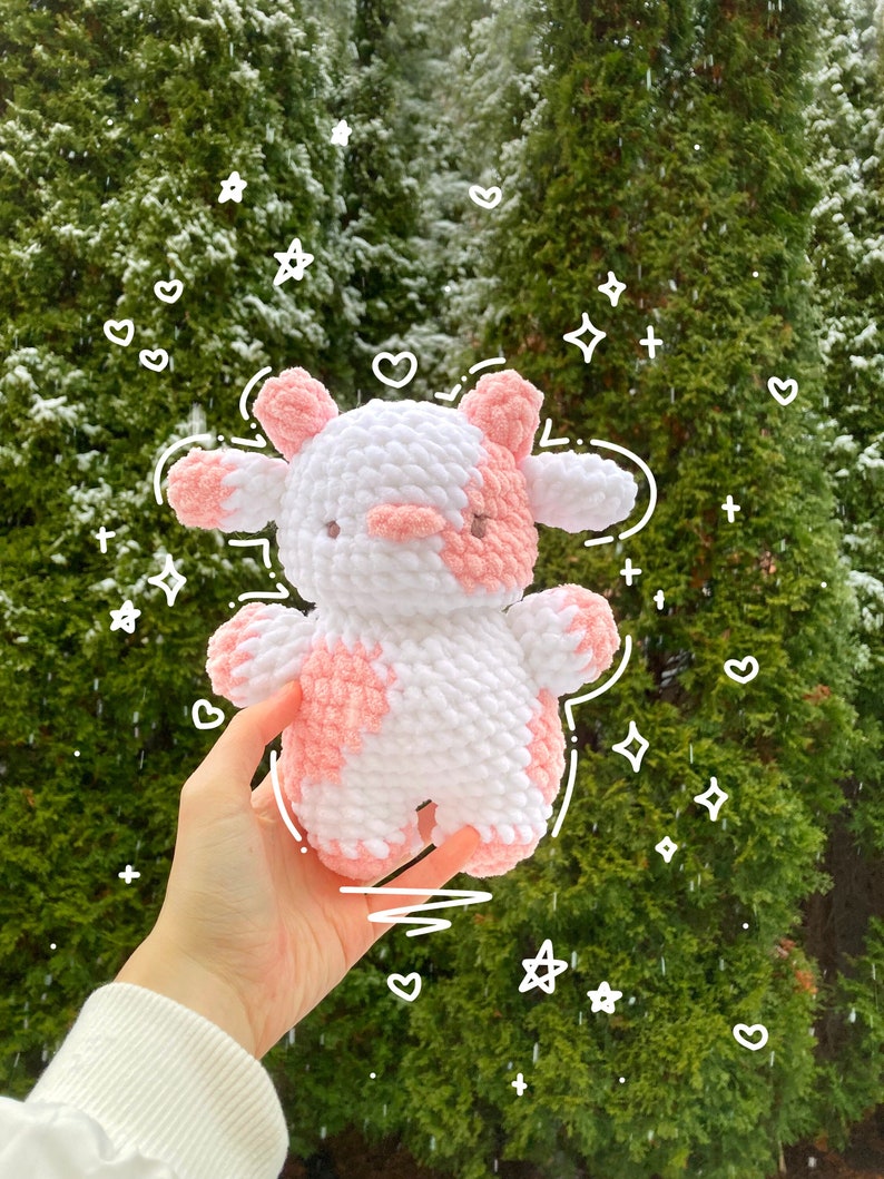 May include: A crocheted pink and white cow plush toy with a fuzzy texture. The toy is being held in a hand against a green background with white snow falling.
