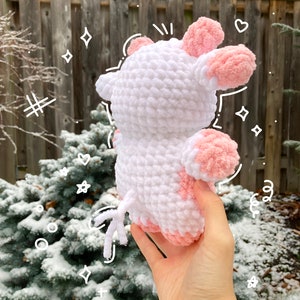May include: A white crocheted plush toy shaped like a cow with pink ears and a pink patch on its side. The toy is being held in a hand against a snowy background.