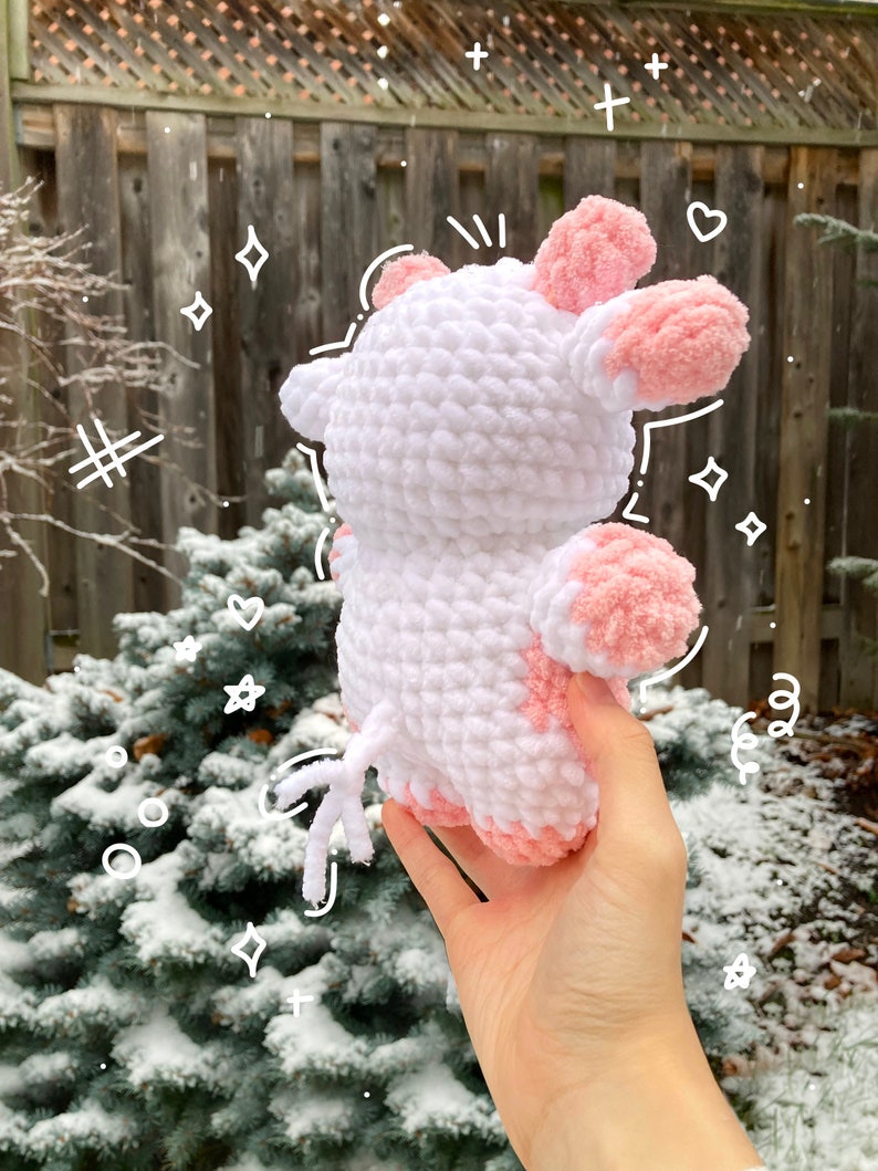 May include: A white crocheted plush toy shaped like a cow with pink ears and a pink patch on its side. The toy is being held in a hand against a snowy background.