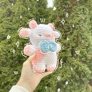 May include: A pink and white crocheted cow plush toy with a blue bow. The toy is being held in a hand against a green background.
