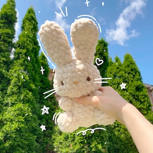 May include: A soft, cream-colored plush bunny rabbit toy with a sweet expression. The bunny is being held up in the air by a hand against a backdrop of green trees and a blue sky.