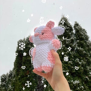 May include: A pink and white crocheted cow plush toy held in a hand against a snowy background of evergreen trees.