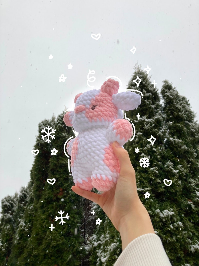 May include: A pink and white crocheted cow plush toy held in a hand against a snowy background of evergreen trees.