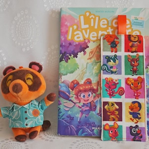 May include: A white fabric bookmark with colourful squares featuring Animal Crossing characters. The bookmark has a bright orange ribbon attached to the top. The bookmark is lying on a white surface next to a book titled "L'ile de l'aventure".