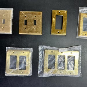 May include: A collection of six rectangular brass light switch plates in various configurations, including single toggle, double toggle, and rocker styles. Some plates are enclosed in clear plastic packaging, while others are exposed.