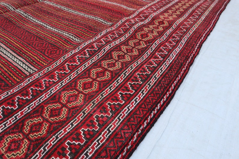 Puede incluir: Primer plano de una alfombra tejida roja, negra y blanca con intrincados patrones geom&eacute;tricos. La alfombra tiene un borde con flecos y est&aacute; doblada por la mitad.