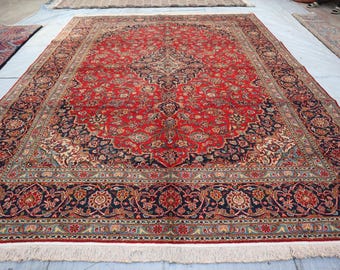 May include: A large, rectangular Persian rug with a central medallion design. The rug features a vibrant red field with intricate floral patterns in navy, cream, and gold. A navy border with floral motifs frames the central design. The rug has a fringed edge.