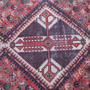 Puede incluir: Primer plano de una alfombra vintage con un patr&oacute;n geom&eacute;trico. La alfombra presenta un fondo rojo con formas de diamante y un dise&ntilde;o central en forma de cruz en rojo, blanco y morado oscuro. La alfombra tiene una apariencia desgastada y envejecida.