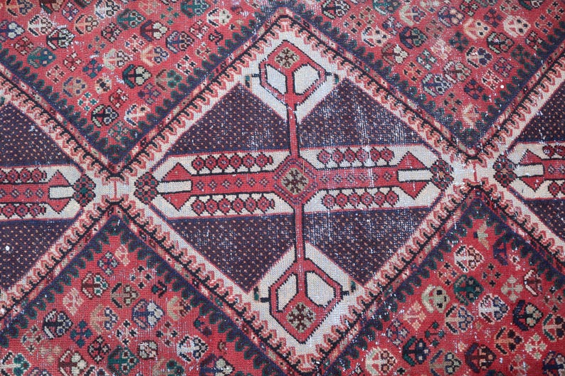 Puede incluir: Primer plano de una alfombra vintage con un patr&oacute;n geom&eacute;trico. La alfombra presenta un fondo rojo con formas de diamante y un dise&ntilde;o central en forma de cruz en rojo, blanco y morado oscuro. La alfombra tiene una apariencia desgastada y envejecida.