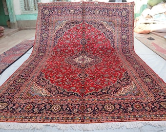 May include: A large, rectangular red rug with an intricate floral pattern and a dark blue border. The rug features a central medallion design and is displayed on a white surface. The rug has a fringed edge.