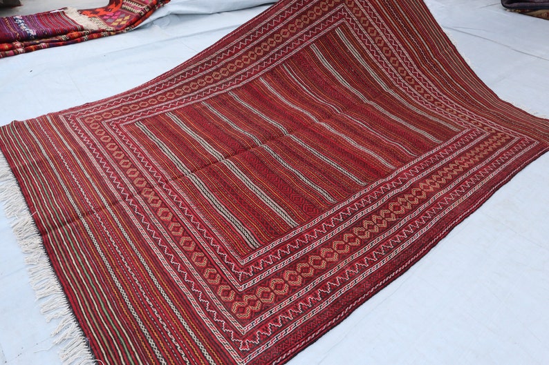 Puede incluir: Una alfombra roja, blanca y negra a rayas con un borde de patr&oacute;n geom&eacute;trico. La alfombra tiene flecos en todos los lados.