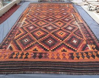 May include: A large, rectangular rug with a geometric diamond pattern in shades of orange, brown, and black. The rug has a border with a repeating design and fringe along the bottom edge. The rug is placed on a blue surface.