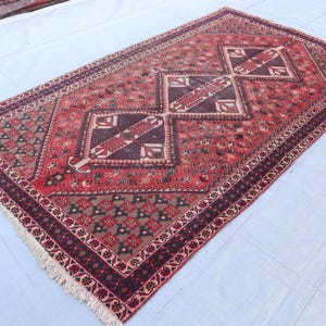 Puede incluir: Una alfombra rectangular anudada a mano, con una combinaci&oacute;n de colores rojo y marr&oacute;n. Presenta tres medallones en forma de diamante en marr&oacute;n oscuro y crema, sobre un fondo rojo. El borde tiene dise&ntilde;os florales y flecos.