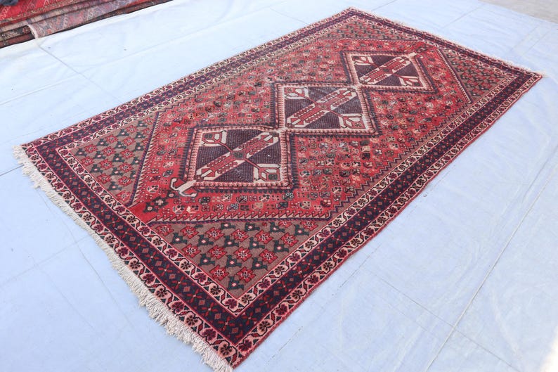 Puede incluir: Una alfombra rectangular anudada a mano, con una combinaci&oacute;n de colores rojo y marr&oacute;n. Presenta tres medallones en forma de diamante en marr&oacute;n oscuro y crema, sobre un fondo rojo. El borde tiene dise&ntilde;os florales y flecos.
