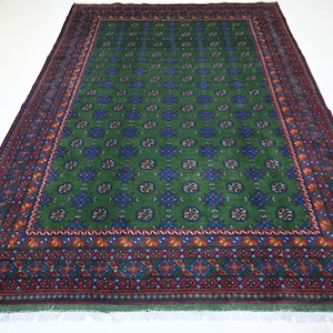 Puede incluir: Una alfombra rectangular verde con un patrón detallado. La alfombra presenta un campo central verde con motivos florales azules y blancos repetidos. Un borde de diseños geométricos rojos, azules y naranjas enmarca la alfombra. La alfombra tiene flecos en los bordes.