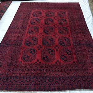 Puede incluir: Una alfombra rectangular con un fondo rojo intenso y un patrón repetitivo de motivos octogonales en azul marino oscuro. La alfombra incluye un borde ancho con formas geométricas intrincadas y flecos en los bordes cortos del tejido.