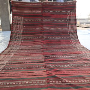 May include: A red, green, and white striped rug with intricate patterns woven into the fabric. The rug is folded in half and being held up by two people.