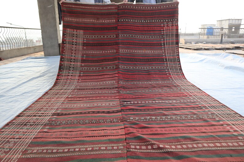 May include: A red, green, and white striped rug with intricate patterns woven into the fabric. The rug is folded in half and being held up by two people.