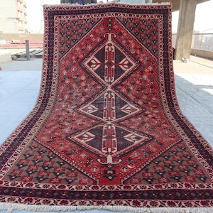 Puede incluir: Una alfombra rectangular grande con un patr&oacute;n de diamantes central en tonos de rojo, marr&oacute;n y crema. La alfombra presenta intrincados dise&ntilde;os geom&eacute;tricos y un borde oscuro con un borde con flecos. La alfombra se exhibe al aire libre.