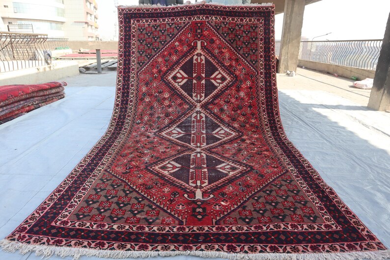 Puede incluir: Una alfombra rectangular grande con un patr&oacute;n de diamantes central en tonos de rojo, marr&oacute;n y crema. La alfombra presenta intrincados dise&ntilde;os geom&eacute;tricos y un borde oscuro con un borde con flecos. La alfombra se exhibe al aire libre.