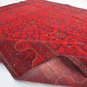 Op de afbeelding: Een rood tapijt met een traditioneel Perzisch ontwerp. Het tapijt heeft een donkerblauwe rand en ingewikkelde patronen over de hele oppervlakte. Het tapijt is dubbelgevouwen, waardoor de achterkant zichtbaar is.