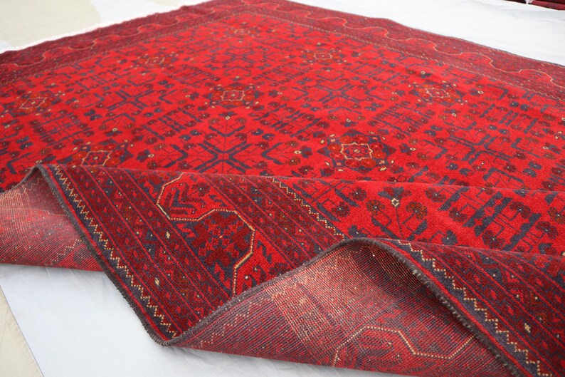 Op de afbeelding: Een rood tapijt met een traditioneel Perzisch ontwerp. Het tapijt heeft een donkerblauwe rand en ingewikkelde patronen over de hele oppervlakte. Het tapijt is dubbelgevouwen, waardoor de achterkant zichtbaar is.