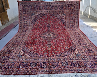 May include: A large, ornate red rug with a central medallion design and intricate floral patterns. The rug features a dark blue border and a cream-coloured fringe. The rug is displayed outdoors, showcasing its size and detail.
