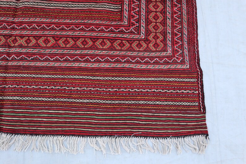 Puede incluir: Una alfombra roja, negra y blanca con flecos. La alfombra tiene un dise&ntilde;o geom&eacute;trico y est&aacute; hecha de lana.