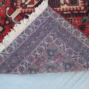 Puede incluir: Una secci&oacute;n de una alfombra estampada roja y beige con un dise&ntilde;o detallado. La alfombra tiene un borde morado oscuro con motivos florales y flecos color crema. La alfombra est&aacute; doblada, revelando la parte inferior.