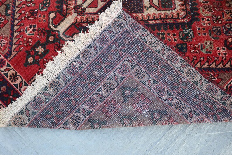Puede incluir: Una secci&oacute;n de una alfombra estampada roja y beige con un dise&ntilde;o detallado. La alfombra tiene un borde morado oscuro con motivos florales y flecos color crema. La alfombra est&aacute; doblada, revelando la parte inferior.