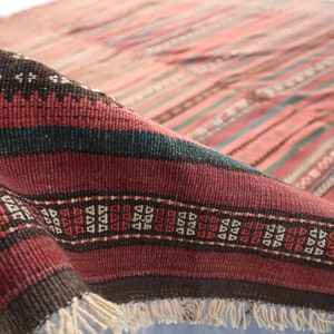 May include: A close-up of a red, brown, and green striped rug with a geometric pattern. The rug has fringe on the bottom edge.