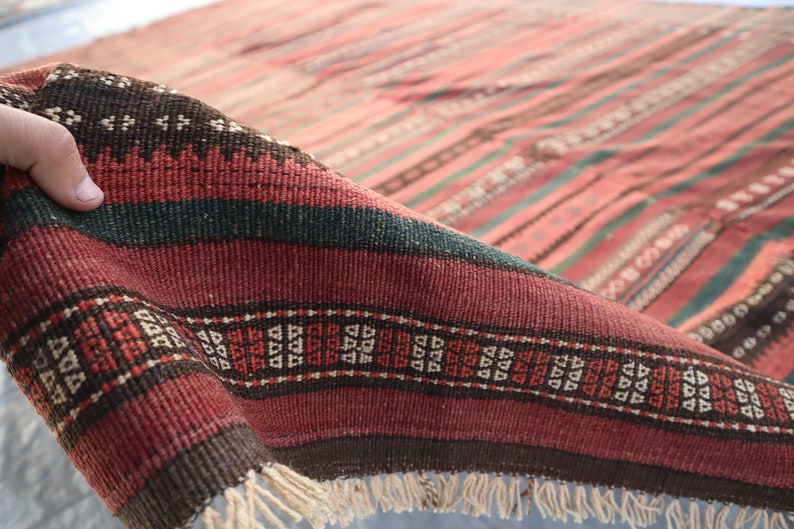 May include: A close-up of a red, brown, and green striped rug with a geometric pattern. The rug has fringe on the bottom edge.