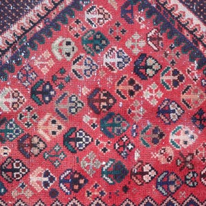 Puede incluir: Primer plano de una alfombra roja vintage con un patr&oacute;n detallado. La alfombra presenta formas geom&eacute;tricas en varios colores, incluyendo azul marino, verde azulado y crema, sobre un fondo rojo descolorido. El dise&ntilde;o incluye motivos de diamantes y flores.