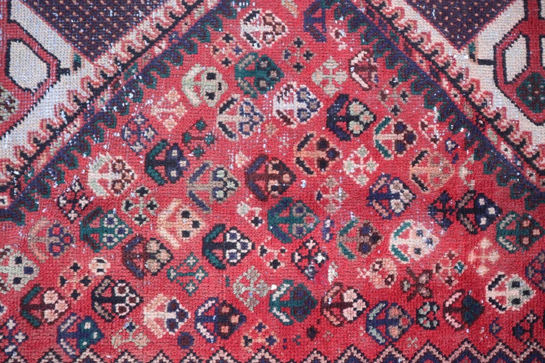 Puede incluir: Primer plano de una alfombra roja vintage con un patr&oacute;n detallado. La alfombra presenta formas geom&eacute;tricas en varios colores, incluyendo azul marino, verde azulado y crema, sobre un fondo rojo descolorido. El dise&ntilde;o incluye motivos de diamantes y flores.