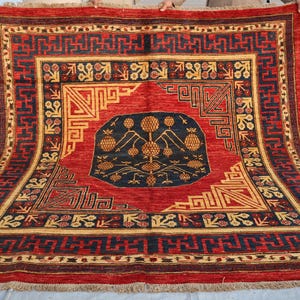 May include: A rectangular, hand-woven rug with a central medallion design. The rug features a red field with a navy blue and gold border. The central design includes a navy blue oval with gold floral motifs and geometric patterns.