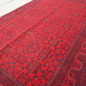 Op de afbeelding: Een rood tapijt met een geometrisch patroon in blauw en goud. Het tapijt heeft een rand met een herhalend ontwerp.